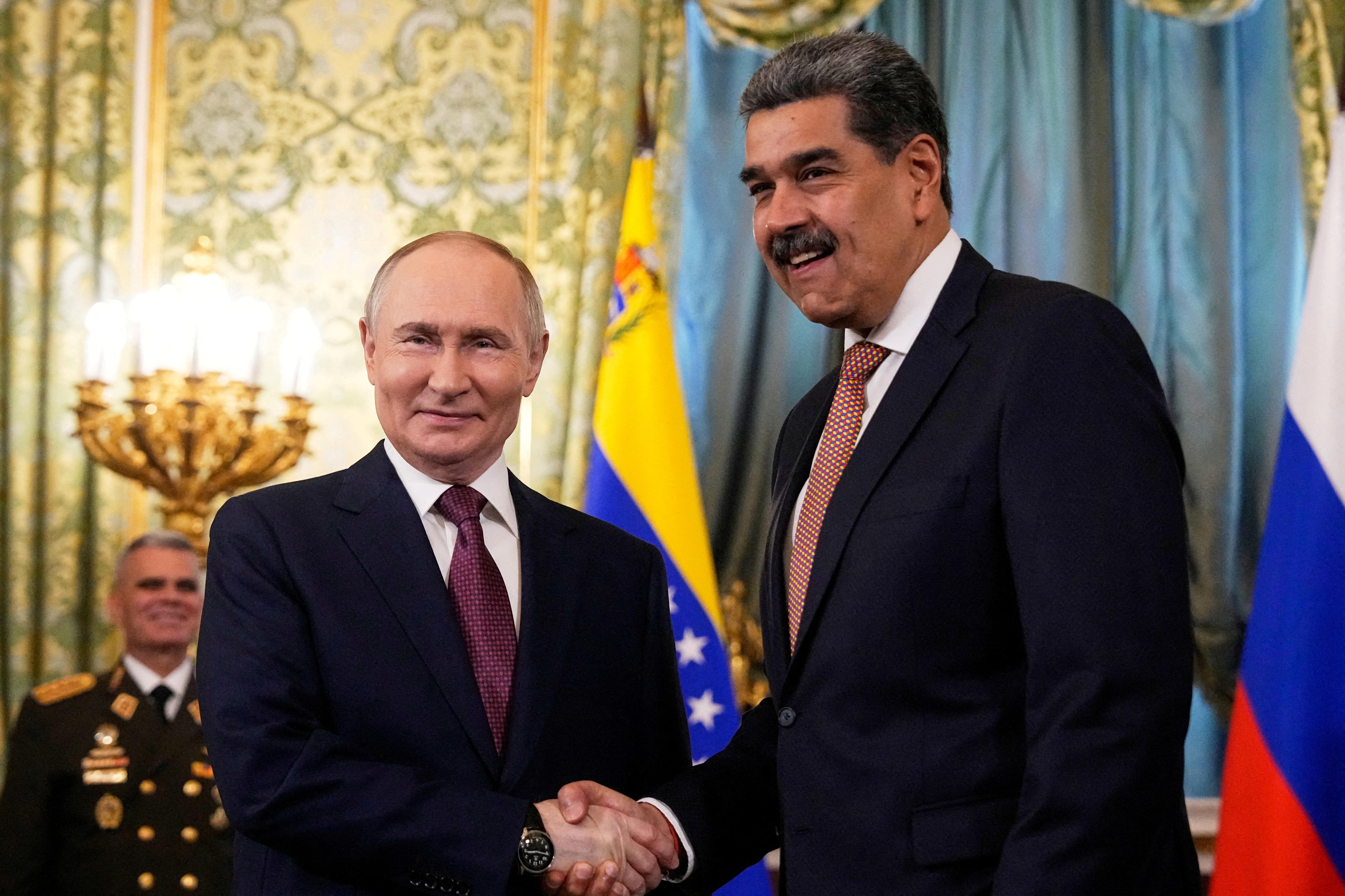 Vladimir Putin and Nicolas Maduro in dark suits shaking hands and smiling in a room with floral wallpaper and national flags