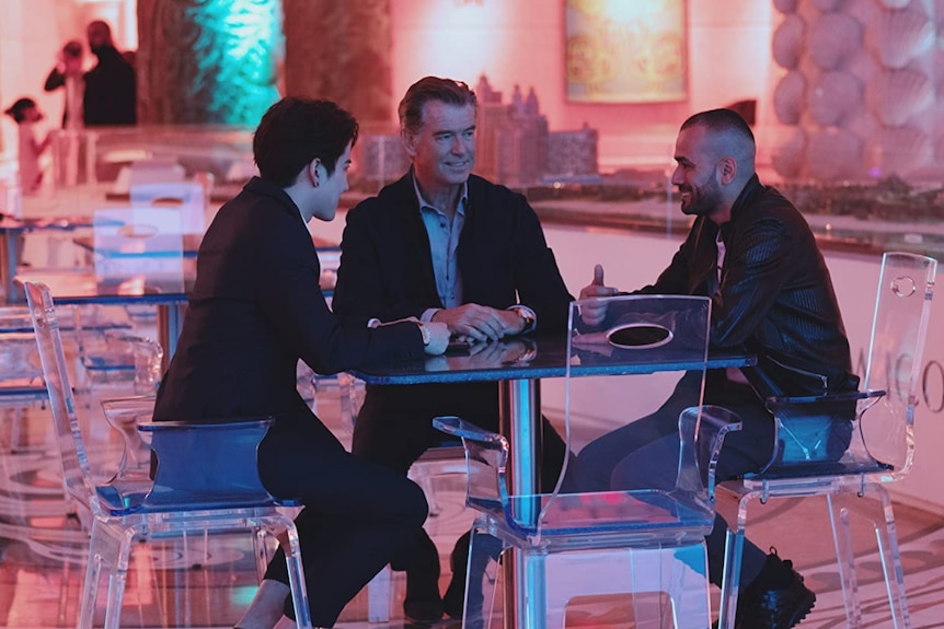 Three male actors sit at a table with clear chairs on a movie set, talking to each other