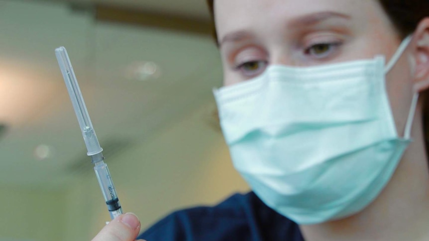 A nurse in a face mask holds and inspects a vaccination needle.