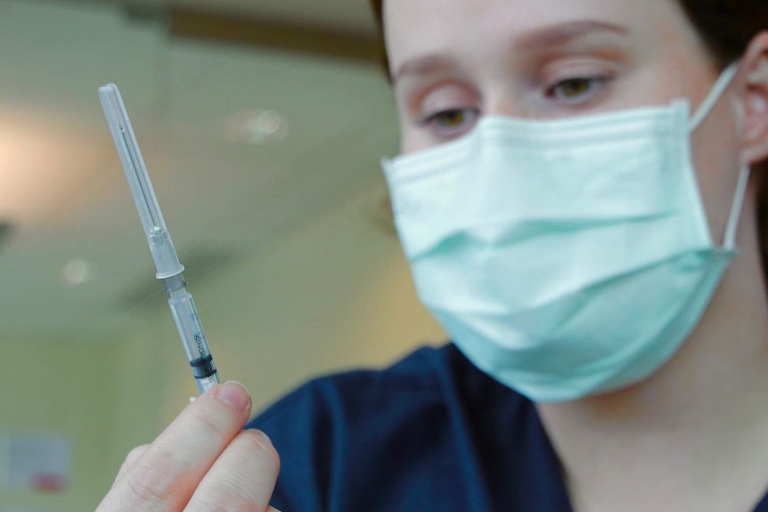 A nurse in a face mask holds and inspects a vaccination needle.