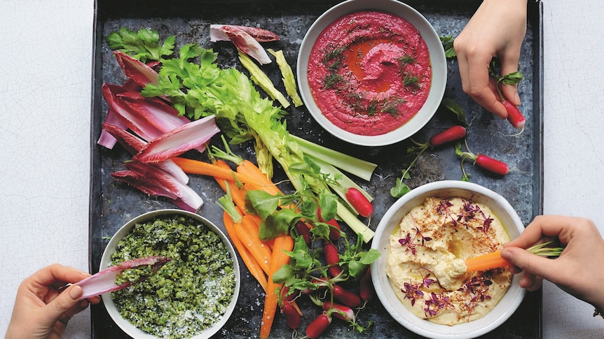 Three dips (hummus, beetroot and pesto) in ramekins on a roasting tray alongside celery and other vegetables.