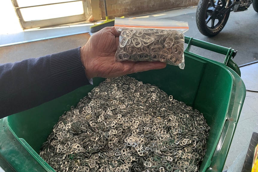 A hand holding a small bag of ring-pulls over an almost full wheelie bin full of ring-pulls.