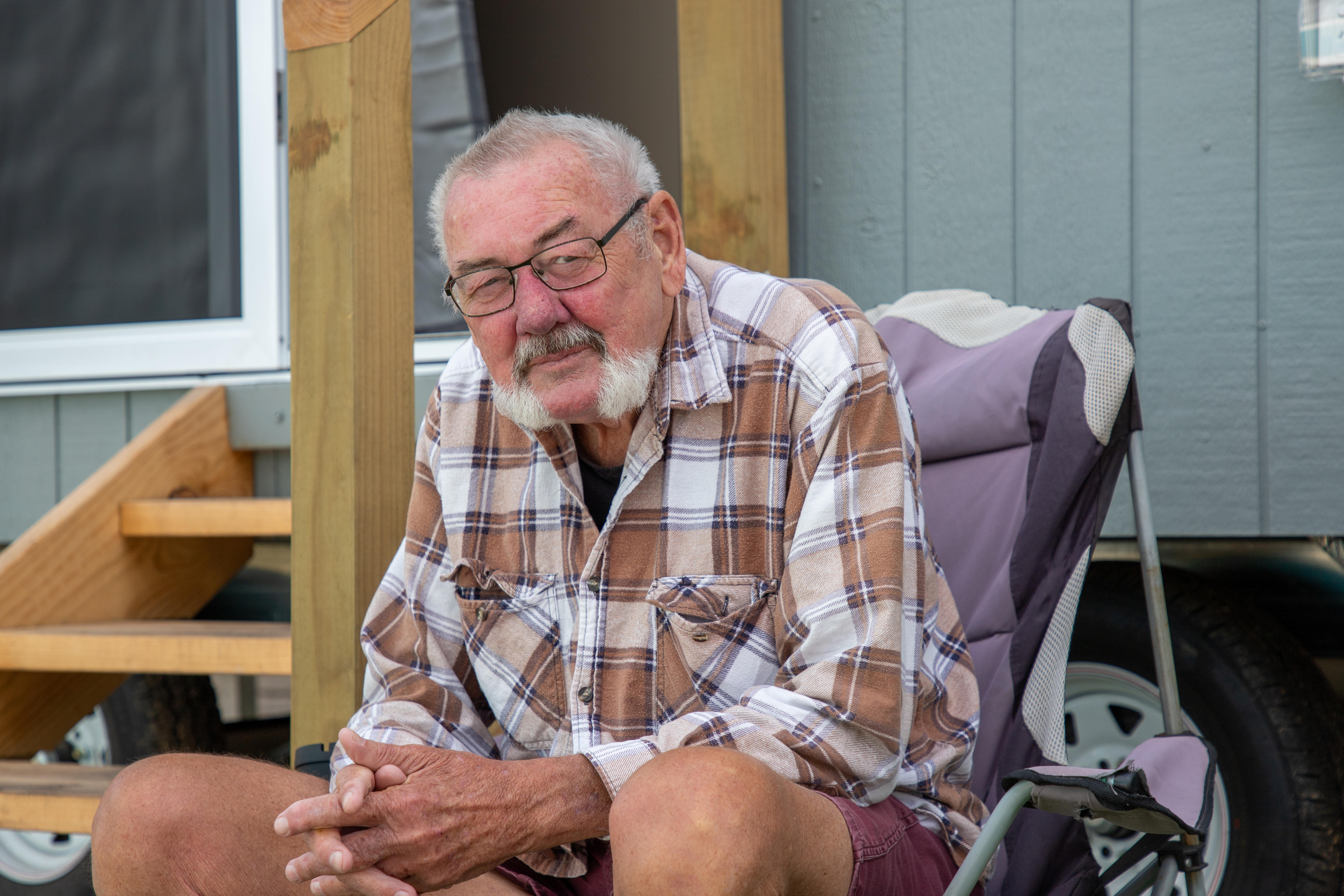 A man with glasses and brown flannelette shirt sits on a purple outdoor chair.