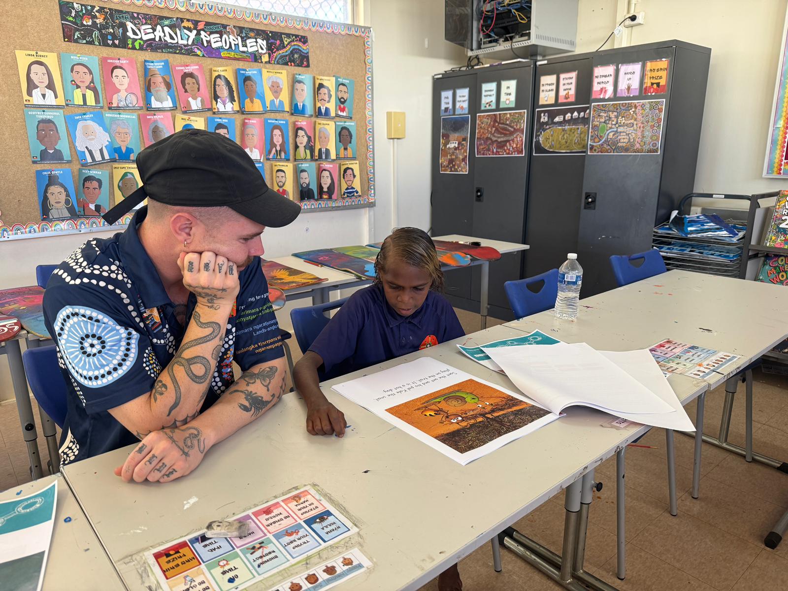 Un hombre con gorra apoya la barbilla en la mano mientras se sienta y lee en un escritorio junto a Brynetta.