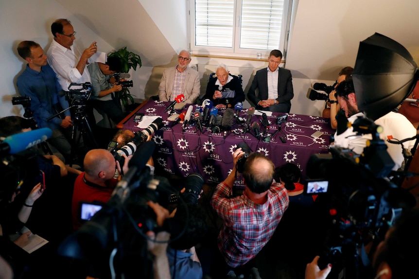 View from above as David Goodall sits at a table flanked by a man and a woman, with the rest of the room packed with media