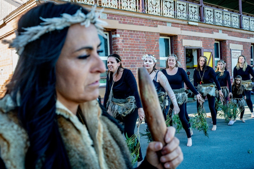 Indigenous woman holds dancing stick as dancers in traditional feathered skirts and headbands follow her.