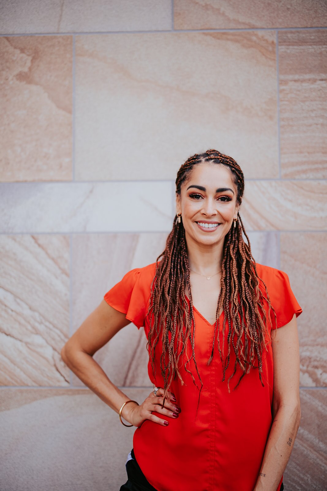 Portrait of a woman of colour with long hair braids. She has a wide smile and is wearing an orange shirt.