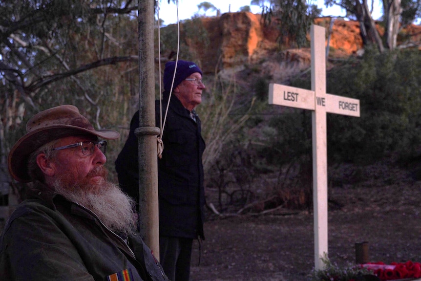 a man with a scruffy beard sitting down, another man in a beanie behind him and a lest we forget cross in the background