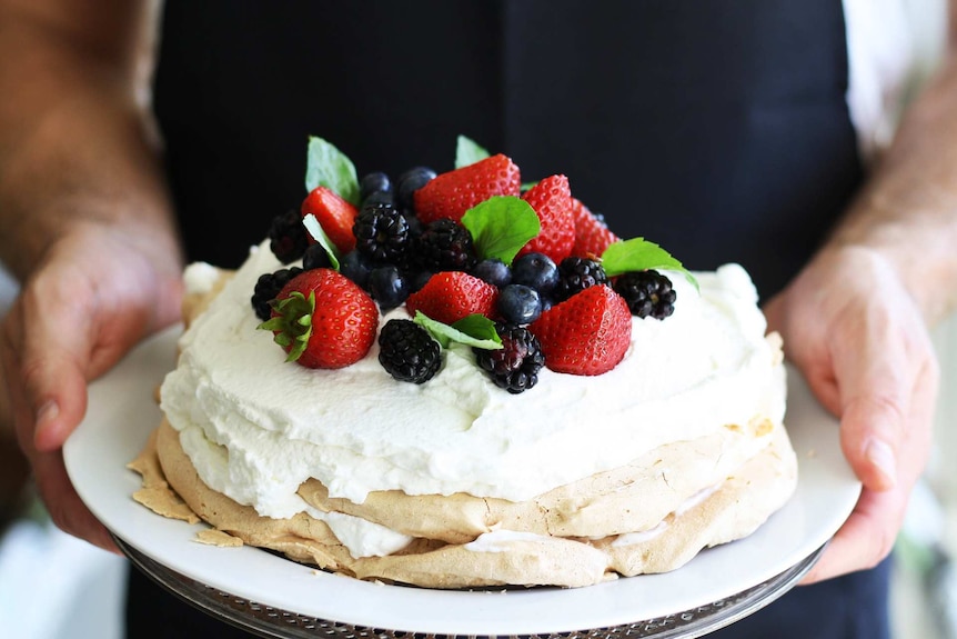 Close up of a man holding a finished pavlova dessert topped with cream, fresh berries and mint.