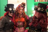 3 women wearing Victorian dressed with red wigs, corset, goggles, top hats and buckles in front of a bookcase lined green wall