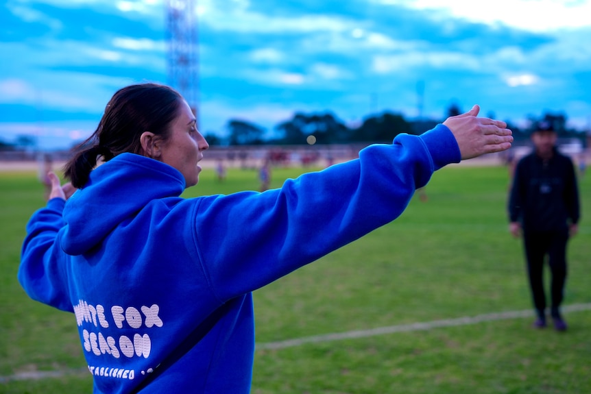 A white woman with brown hair wearing a blue jumper stretching out her arms on a football field. 