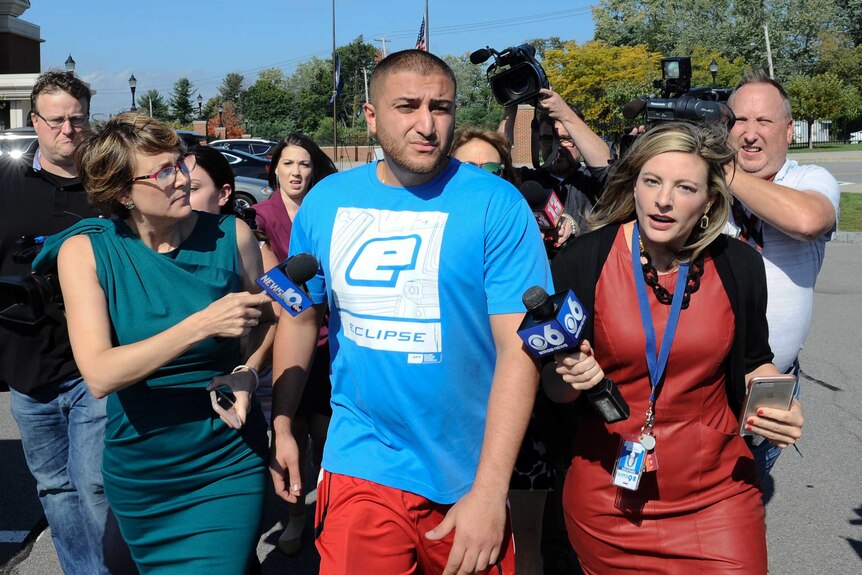 Shahyer Hussain, wearing a blue t-shirt and red pants, mid-stride, outside, with a media pack surrounding him with microphones.