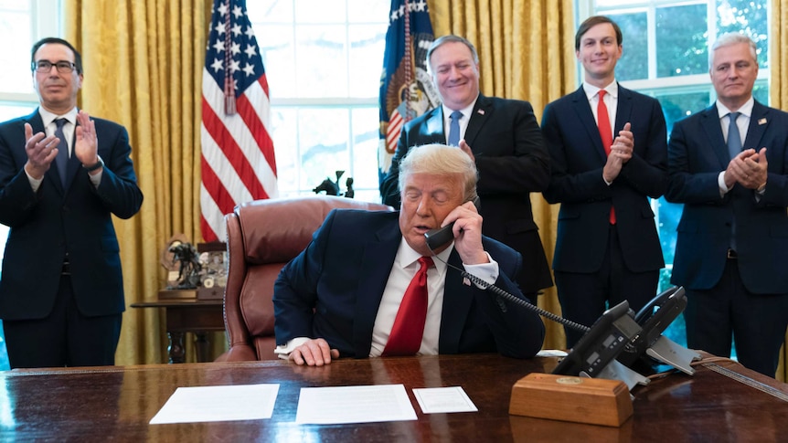 A man in a suit speaks on a phone while four smiling supporters stand behind and clap.