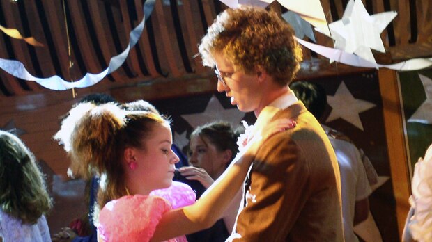 A girl with a pink dress and high pony tail dances awkwardly with a blonde, curly-haired boy in a brown suit.