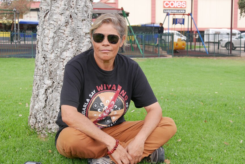 A woman with short grey hair sitting with her legs crossed on the lawn wearing a black shirt, sunglasses and tanned pants. 