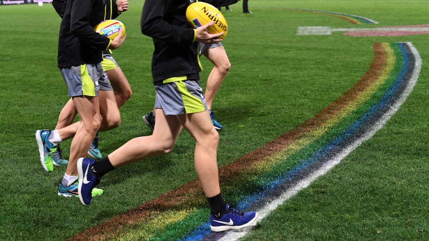 Players, holding footballs, run over a rainbow pattern on a sporting field.