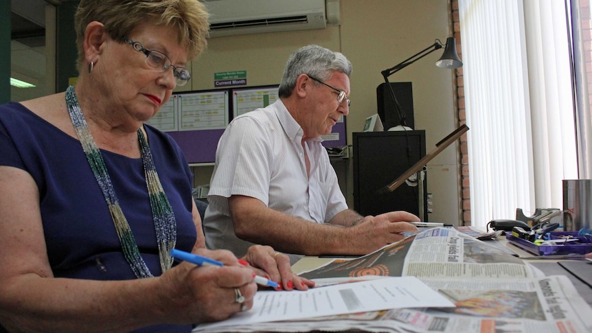 A woman and a man sitting at a desk making notes as they read a newspaper