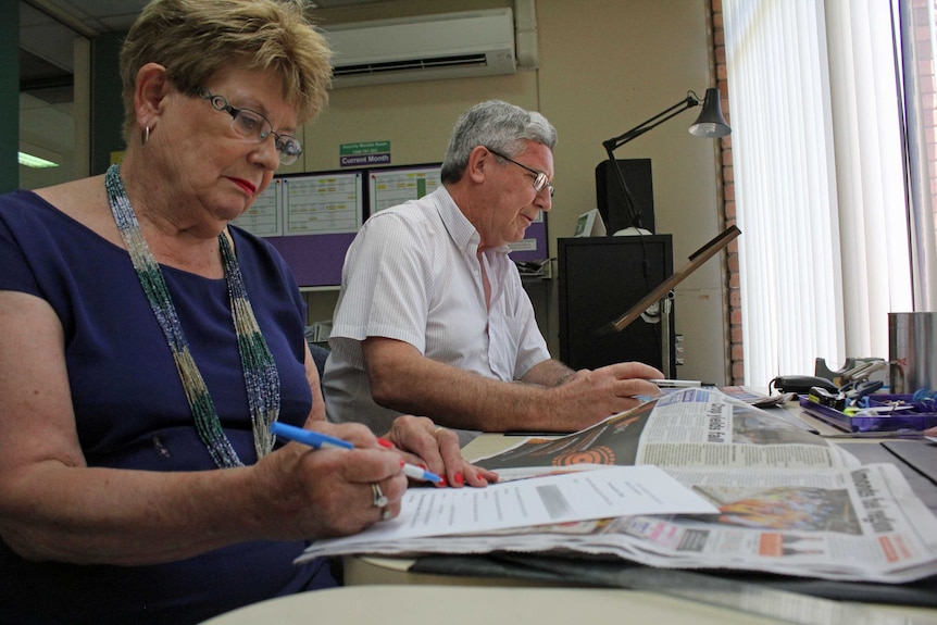 A woman and a man sitting at a desk making notes as they read a newspaper