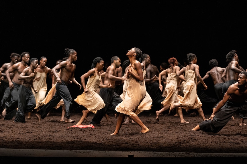 A group of African dancers perform on stage. The stage is covered in a thick layer of dirt. Dancers appear to be mid-run.