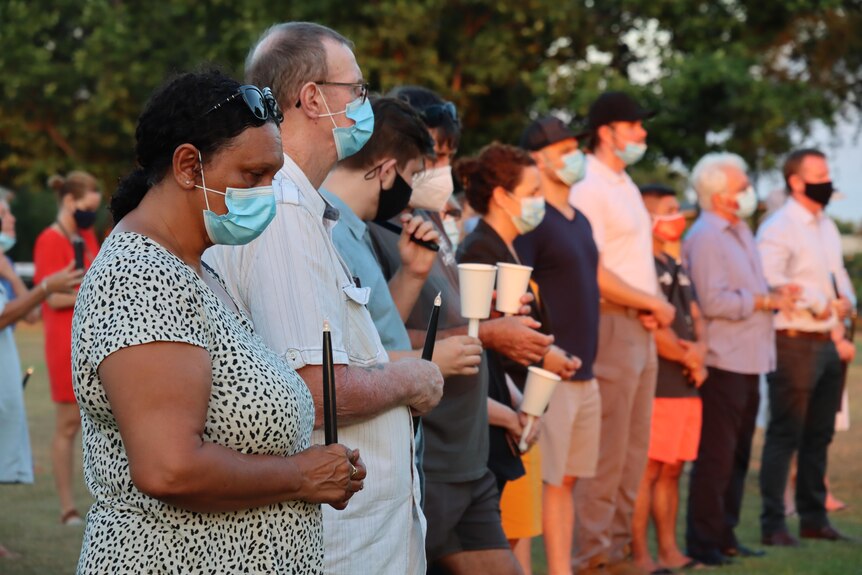 A crowd of people wearing face masks hold candles and appear solemn. A woman in the foreground has black hair.