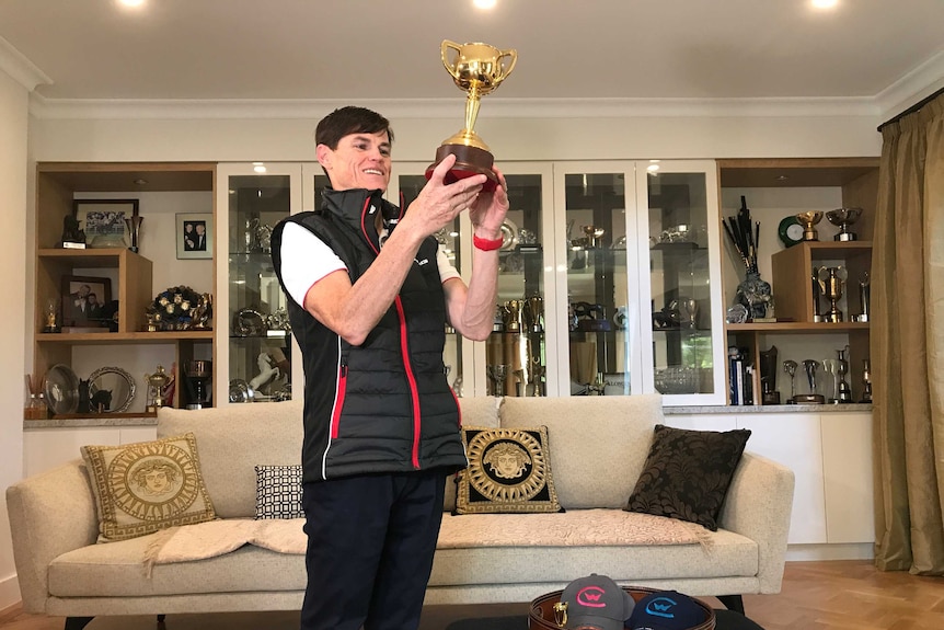 Craig Williams holds his cup in a living room with several trophies and cups on shelves behind him.