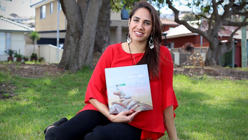 Kirli Saunders sits down on grass, holding her book.