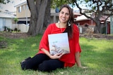 Kirli Saunders sits down on grass, holding her book.