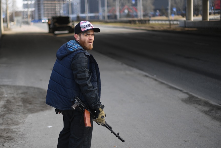 a man with a rifle dressed in a jacket and a hat looks over his shoulder at the camera as he stands on a street near an overpass