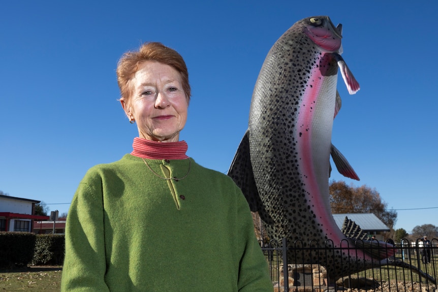 An older woman smiling beside a statue of a big trout.