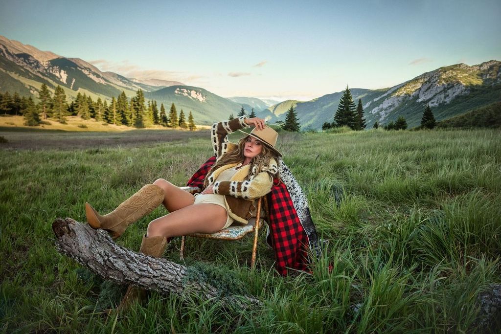 Country artist Shania Twain lounges in a chair in a mountain range in a jumpsuit, cowboy boots, and cowboy hat.