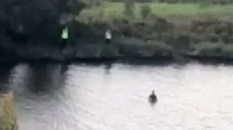 A screenshot of two police officers on a riverbank and a boy in the water.
