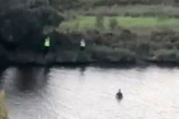 A screenshot of two police officers on a riverbank and a boy in the water.