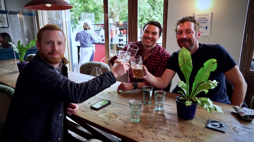 Three men cheers with beer glasses across a bar table.