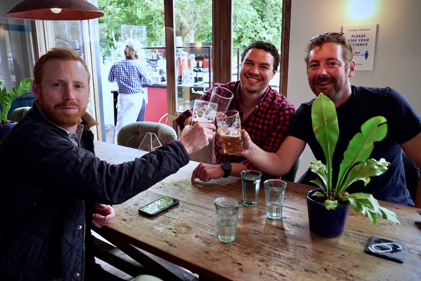 Three men cheers with beer glasses across a bar table.