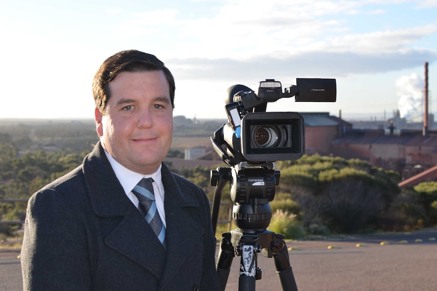 A man wearing a suit in his 20s stands with a video camera at sunset with the steelworks in the background.