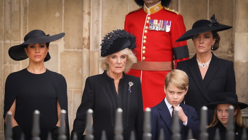 Meghan, Camilla and Princess Kate stand on a balcony wearing black dresses and hats while Prince George stands beside them.