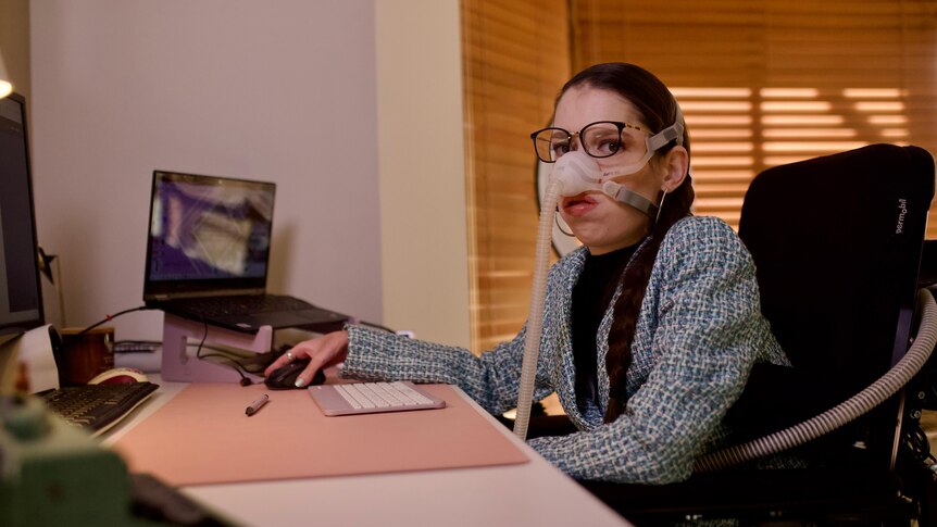 A lady with a ventilator sits at a computer while looking at the camera