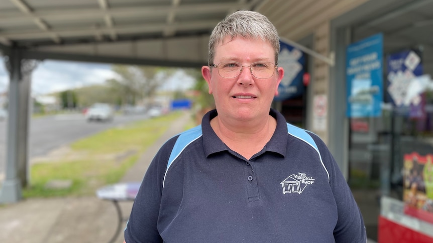 A woman with short hair and a blue shirt stands outside a general store.