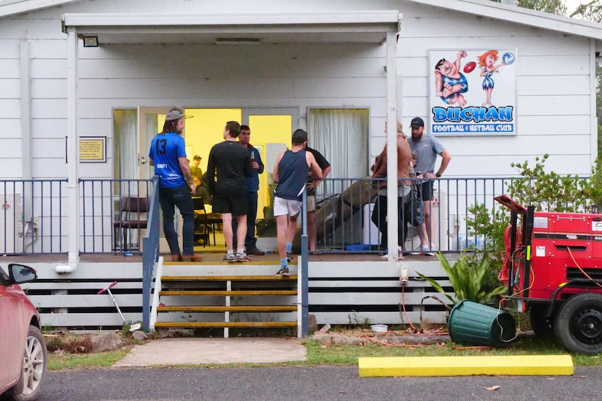 People gathered on a deck in front of a white building, drinking beers.