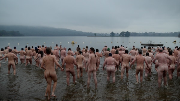 A large number of naked people run or walk into a grey lake, fog hanging in the distance. The camera is positioned behind them. 