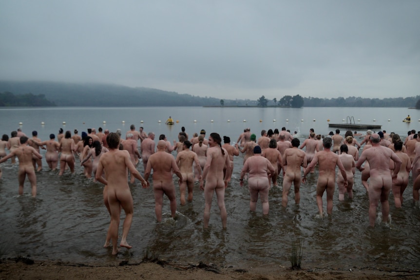 A large number of naked people run or walk into a grey lake, fog hanging in the distance. The camera is positioned behind them. 