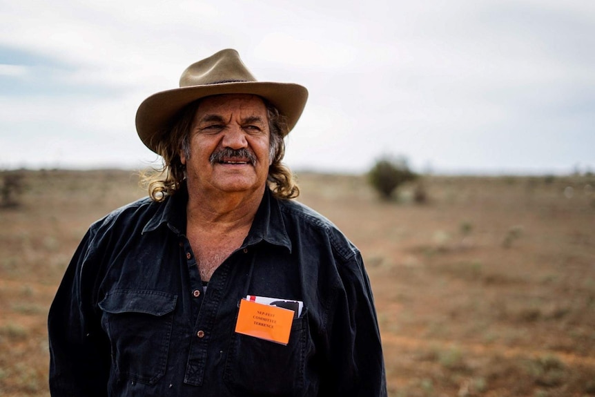 An Indigenous man in a blue work shirt and hat, standing in a dry paddock.
