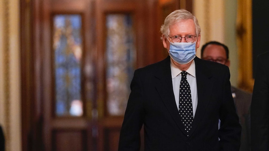 A man with grey hair wearing a dark suit and a mask walks away from doors.