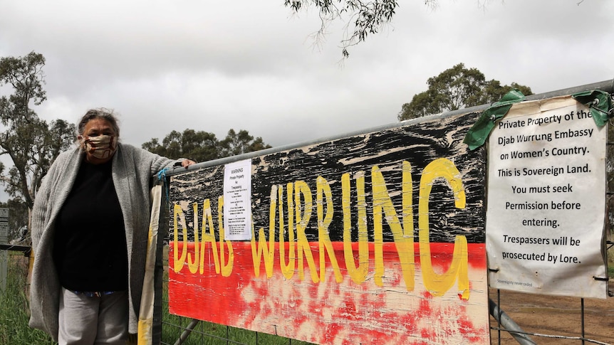 A woman standing next to a gate with signs emblazoned on it including 'Djab Wurrung'.