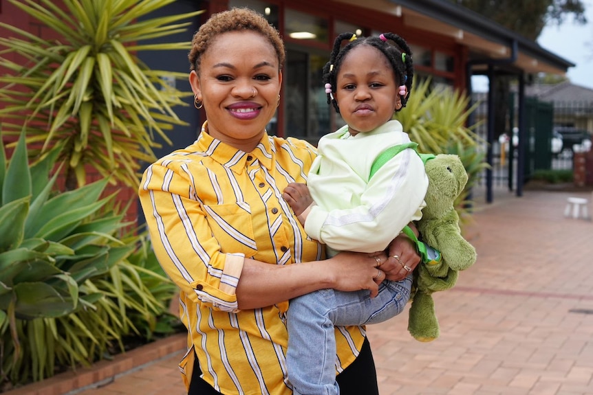 A woman holding her young daughter outside a building with plants nearby