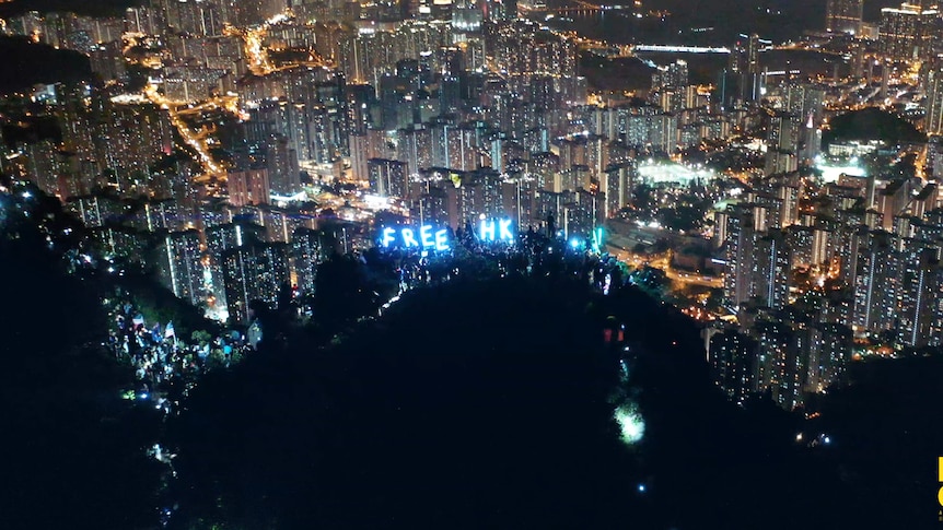 An aerial shot shows the city of Hong Kong by night. Among the city lights is a message that reads FREE HK