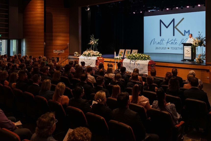 People listen to a priest speak in an auditorium with two caskets.