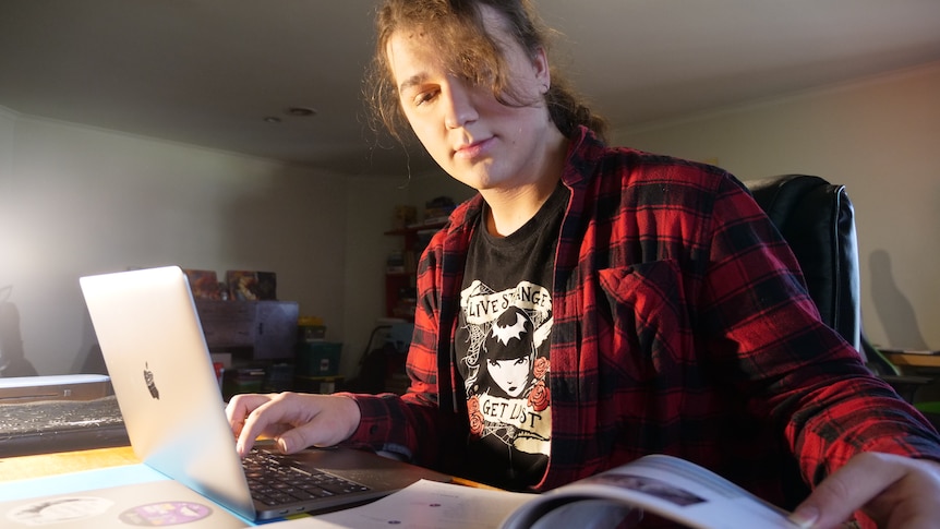 A woman in a flannel shirt looking through a school textbook while also typing on her laptop.
