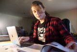 A woman in a flannel shirt looking through a school textbook while also typing on her laptop.