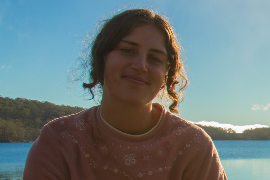 A young woman wearing a pink jumper sits in front of a lake.